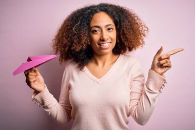 Afro saçlı, pembe arka planda kağıt uçak tutan Afro-Amerikalı genç bir kadın. Elleri ve parmakları kenarı işaret ederek çok mutlu.