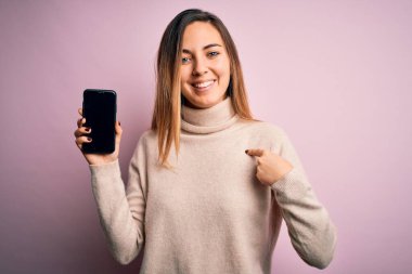 Genç, güzel esmer kadın elinde akıllı telefonuyla pembe arka planda kendini gösteren sürpriz yüzüyle ekranı gösteriyor.