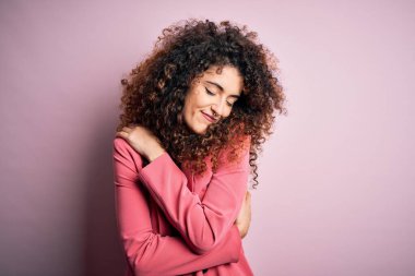 Kıvırcık saçlı ve piercingli genç ve güzel bir iş kadını zarif bir ceket giyiyor. Kendine sarılıyor, mutlu ve pozitif, kendinden emin gülümsüyor. Sevgi ve özgüven.