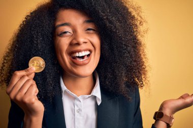 Afro saçlı, sarı arka planda bitcoin tutan Afro-Amerikalı genç bir iş kadını çok mutlu ve heyecanlı, kazanan yüz ifadesi zaferi büyük bir gülümsemeyle kutluyor ve ellerini kaldırıyor.