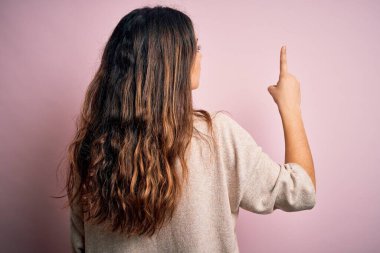 Genç, güzel, esmer bir kadın pembe arka planda dikilip parmak eliyle öne doğru poz veriyor.
