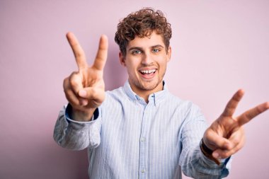 Sarışın, kıvırcık saçlı, beyaz arka planda çizgili tişört giyen, iki elini de zafer işareti yaparken gösteren ve dili dışarı çıkmış genç, yakışıklı bir adam. İki numara..