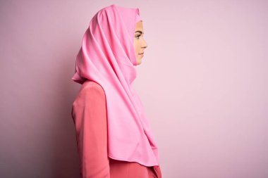 İslami tesettüre bürünmüş genç ve güzel bir kız pembe arka planda dikiliyor, yan yana bakıyor, rahat bir görünüm takınıyor ve kendine güvenen bir gülümsemeyle gülümsüyor..