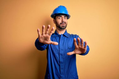 Sakallı, mavi üniformalı ve sarı arka planda güvenlik kaskı takan tamirci. Korkudan dehşete düşmüş ve korkmuş. El hareketlerini durdur, şok içinde bağır. Panik konsepti.