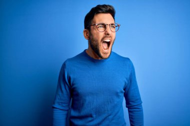 Sakallı, sıradan süveter giyen, mavi arka planda gözlük takan sinirli ve öfkeli sinirli sinirli sinirli bağıran öfkeli genç bir adam. Öfke ve agresif kavram.