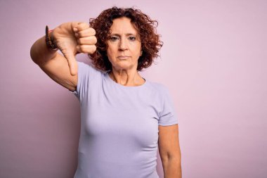 Orta yaşlı güzel kıvırcık saçlı kadın pembe arka plan üzerine günlük tişört giyip mutsuz ve kızgın görünüyor. Reddedilmiş ve olumsuz jestler yapıyor. Kötü ifade.
