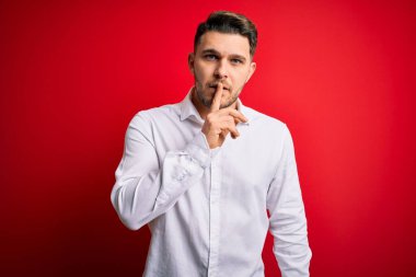 Mavi gözlü, zarif gömlekli genç bir iş adamı kırmızı izole arka planda dikiliyor ve dudakları parmaklarıyla sessiz olmak istiyor. Sessizlik ve gizli konsept.