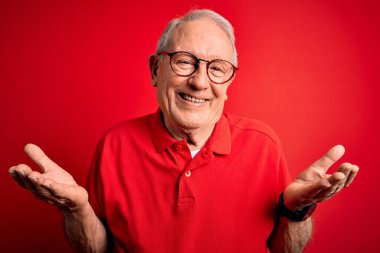 Gri saçlı, gözlüklü ve kırmızı arka planda rahat tişörtlü, kafası karışık, elleri ve kolları havada bir adam. Şüphe kavramı.