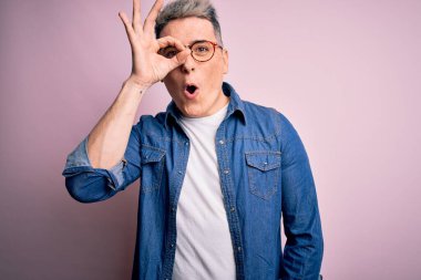 Pembe izole edilmiş arka planda gözlüklü ve kot ceketli genç, yakışıklı ve modern bir adam şaşırmış yüz ve gözlerle parmaklarından bakıyor. İnançsız ifade.