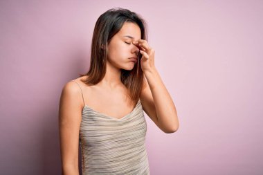 Genç ve güzel esmer kız, izole edilmiş pembe arka plan üzerinde günlük çizgili tişört giyiyor. Burnu ve gözleri ovuşturuyor, yorgunluk ve baş ağrısı hissediyor. Stres ve hüsran kavramı.