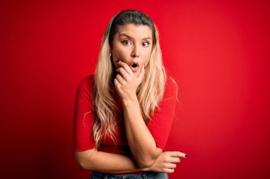 İzole edilmiş kırmızı arka plan üzerinde duran gündelik tişört giyen genç ve güzel sarışın kadın elleri çenesinde olan inançsızlıktan, şaşkınlıktan ve şaşkınlıktan büyülenmiş görünüyor.