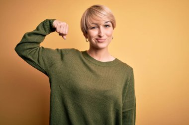 Sarışın, modern kısa saçlı, sarı arka planda rahat süveter giyen güçlü kol kası olan, kendinden emin ve gururlu bir kadın.