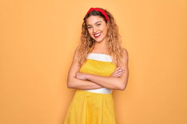 Sarı arka planda dikey duran mavi gözlü güzel sarışın kadın kameraya bakarken kollarını çapraz yapıp gülümsüyor. Pozitif kişi.