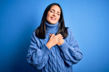 Mavi gözlü genç esmer kadın balıkçı yaka kazak giyiyor elleri göğsünde, gözleri kapalı gülümsüyor ve yüzünde minnet ifadesi var. Sağlık kavramı.