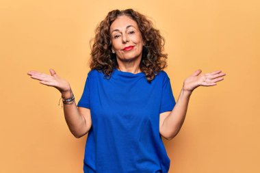 Orta yaşlı, rahat tişört giyen güzel bir kadın izole edilmiş sarı arka planda duruyor. Hiçbir şeyden habersiz, kolları açık, hiçbir fikri yok ve şüpheli bir yüzü var..