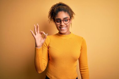 Sarı arka planda süveter ve gözlük takan güzel Afro-Amerikan kızı gülümseyerek el ve parmaklarıyla tamam işareti yapıyor. Başarılı ifade.