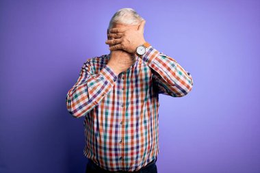 Üst düzey yakışıklı beyaz adam, izole edilmiş mor arka plan üzerine gündelik bir gömlek giyiyor gözleri ve ağzı kapalı, şaşırmış ve şok olmuş. Duygularını gizliyor.