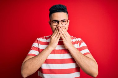 Genç, çizgili tişört ve gözlük takan yakışıklı bir adam izole edilmiş kırmızı arka planda gülüyor ve utanıyor kıkırdıyor ağzı ellerle, dedikoduyla ve skandal konseptiyle kaplıyor.