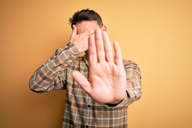 Genç, yakışıklı bir adam, günlük gömleğini giymiş, izole edilmiş sarı arka plan üzerinde duruyor, gözleri ellerle örtüyor ve üzgün ve korku ifadeleriyle jestleri durduruyor. Utanç verici ve olumsuz bir kavram..