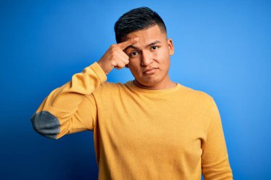 İzole edilmiş mavi arka plan üzerine sarı kazak giyen genç, yakışıklı latin adam alnında sivilceden mutsuz olduğunu gösteriyor, siyah noktalı çirkin bir enfeksiyon. Sivilce ve deri problemi