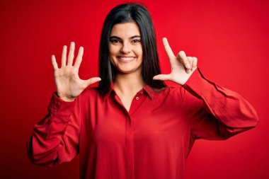 Genç, güzel, esmer, rahat gömlekli bir kadın kırmızı arka planda dikiliyor ve yedinci parmaklarıyla işaret ederken kendinden emin ve mutlu gülümsüyor..