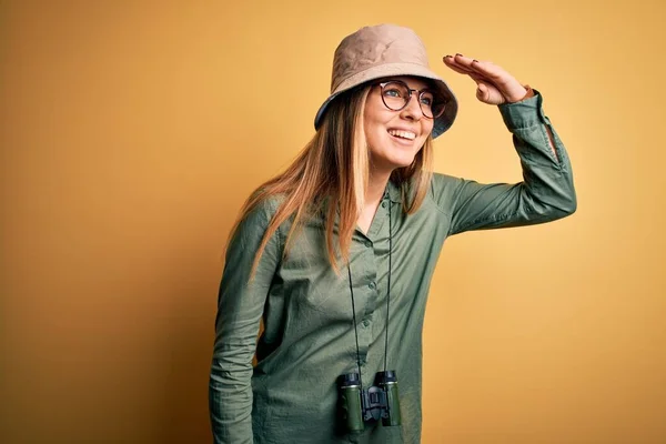 Beautiful blonde explorer woman with blue eyes wearing hat and glasses using binoculars very happy and smiling looking far away with hand over head. Searching concept.