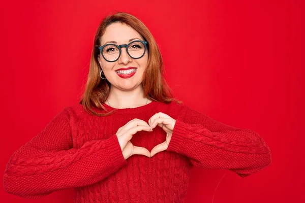 赤い背景にカジュアルなセーターと眼鏡を身に着けている若い美しい赤毛の女性は 手で心のシンボルと形状を示す愛に笑みを浮かべて ロマン主義 — ストック写真