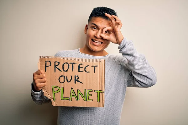 Young handsome activist latin man holding banner asking to protect our planet with happy face smiling doing ok sign with hand on eye looking through fingers