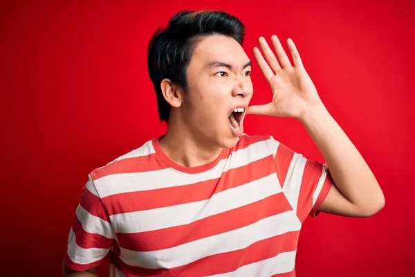 若いハンサムな中国人男性のカジュアルな縞模様のTシャツの赤い背景に叫んで立って大声で手を口の上に手で側に叫んでいる コミュニケーションの概念 — ストック写真