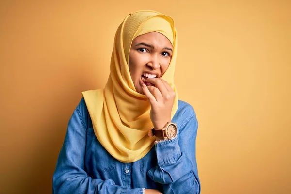 黄色の背景の上にアラブの伝統的なヒジャーブを身に着けている巻き毛を持つ若い美しい女性は 口の上に爪をかむ手で強調し 緊張している 不安の問題 — ストック写真