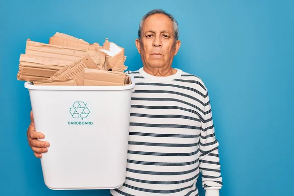 Senior man recycling holding trash can with cardboard to recycle over blue background with a confident expression on smart face thinking serious
