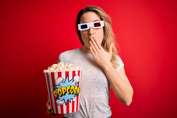 Young beautiful blonde woman watching movie using 3d glasses eating popcorn snack cover mouth with hand shocked with shame for mistake, expression of fear, scared in silence, secret concept