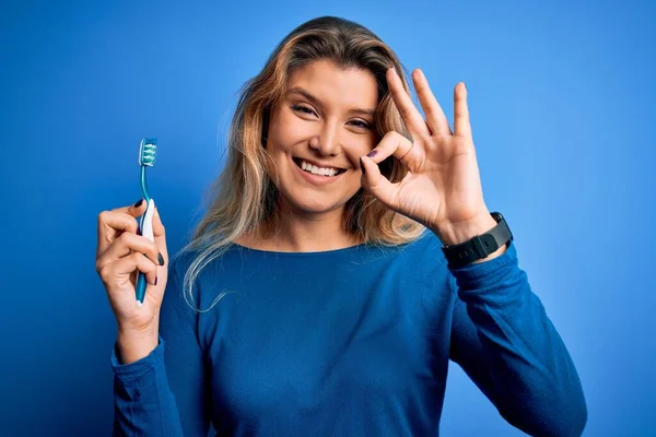 年轻美丽的金发女人用刷子刷着孤立的蓝色背景 用手指做着好的手势 很好的符号 — 图库照片
