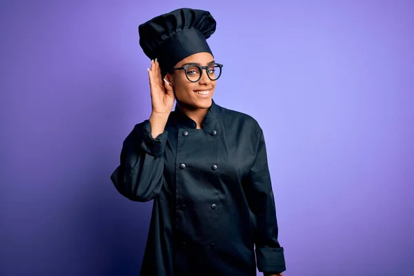 Young african american chef girl wearing cooker uniform and hat over purple background smiling with hand over ear listening an hearing to rumor or gossip. Deafness concept.