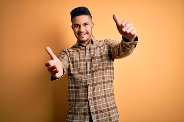 Young handsome man wearing casual shirt standing over isolated yellow background looking at the camera smiling with open arms for hug. Cheerful expression embracing happiness.