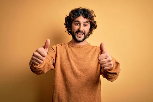 黄色の背景の上に立ってカジュアルなセーターを着てひげを持つ若いハンサムな男は 手で積極的なジェスチャーを行うことを承認し 親指を笑顔と成功のために幸せ 勝者のジェスチャー — ストック写真