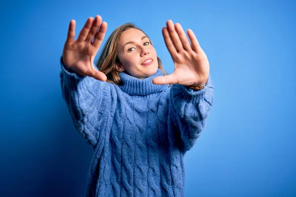 Young beautiful blonde woman wearing casual turtleneck sweater over blue background doing frame using hands palms and fingers, camera perspective