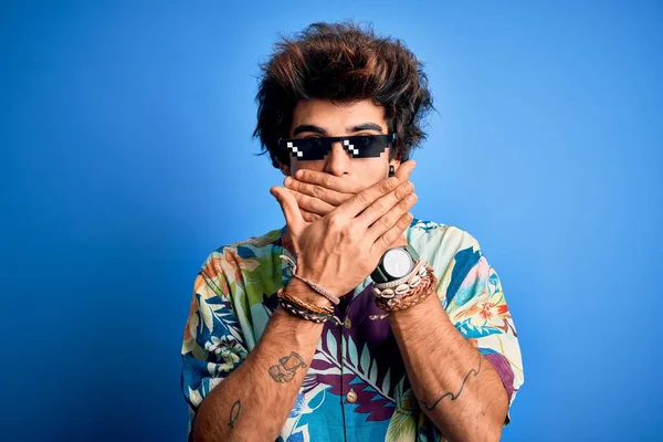 Young handsome man wearing thug life glasses standing over isolated blue background shocked covering mouth with hands for mistake. Secret concept.