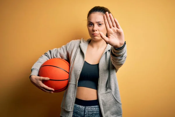 若いです美しいですブルネットスポーツ女性を保持していますバスケットボールボール以上黄色の背景でオープン手を行います停止標識で真剣で自信を持って式 防衛ジェスチャー — ストック写真