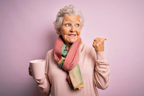 シニア美しいです白髪の女性飲むコーヒーのマグカップ上隔離されたピンクの背景を指すと幸せな顔の笑顔で親指で側面まで表示 — ストック写真