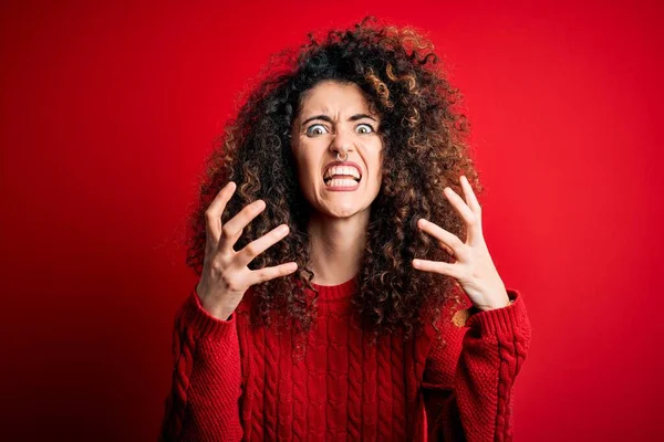 Young beautiful woman with curly hair and piercing wearing casual red sweater Shouting frustrated with rage, hands trying to strangle, yelling mad