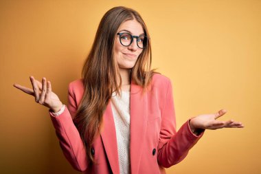 Ceket ve gözlük takan genç, güzel, kızıl bir kadın izole edilmiş sarı arka plan hakkında hiçbir şey bilmiyor ve elleri ve kolları havada, kafası karışmış bir ifade takınıyor. Şüphe kavramı.