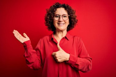 Orta yaşlı, kıvırcık saçlı, rahat gömlekli, kırmızı arka planda gözlüklü güzel bir kadın. Avuç içini gösteriyor ve baş parmaklarıyla hareket ediyor. Mutlu ve neşeli gülümsüyor.