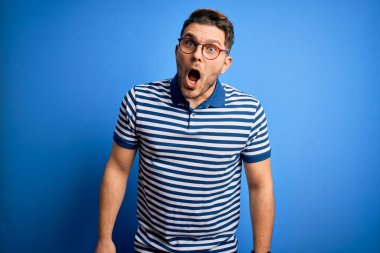 Mavi gözlü, gözlüklü, mavi arka planda sıradan çizgili tişörtlü, korkmuş ve şok olmuş genç bir adam..