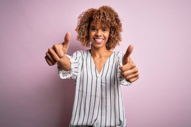 Kıvırcık saçlı güzel Afro-Amerikalı kadın pembe arka planda çizgili tişört giyiyor. Elleriyle olumlu jest yapıyor, gülümsüyor ve başarı için mutlu. Kazanan jest.