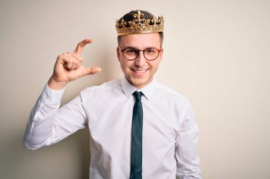 İzole edilmiş arka planda altın taç takan yakışıklı beyaz iş adamı gülümsüyor ve el hareketleriyle küçük boyda işaret yapıyor parmaklarıyla ve kamerayla bakıyor. Ölçü kavramı.