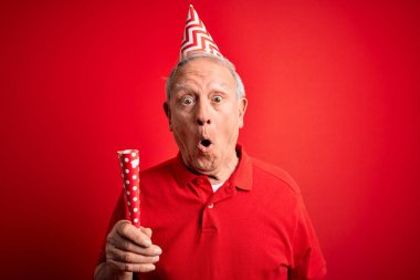 Yaşlı gri saçlı adam doğum günü şapkası takıyor ve kırmızı arka planda parti trompeti tutuyor. Şaşırmış bir yüzle korkmuş, korku ifadesinden korkmuş ve heyecanlanmış.