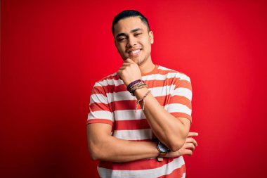 İzole edilmiş kırmızı arka plan üzerinde duran sıradan çizgili tişört giyen genç Brezilyalı adam kameraya kollarını kavuşturup gülümseyerek ve çenesini kaldırarak bakıyor. Olumlu düşünüyorum..