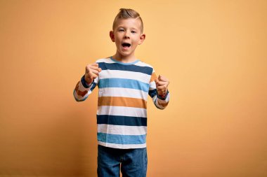 Sarı arka planda mavi çizgili tişört giyen, mavi gözlü küçük beyaz çocuk kollarını kaldırarak ve açık gözlerle başarıyı kutluyor ve şaşırıyor. Kazanan konsept.