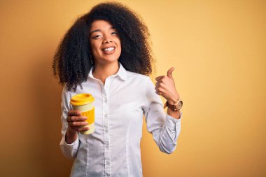 Afro saçlı, kahve fincanından kahve içen, gülümseyen, işaret veren, parmaklarıyla başparmağını kaldıran Afro-Amerikalı genç bir iş kadını.